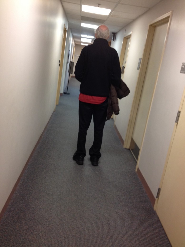 Back view of older man walking down medical hallway with a woman's coat under his arm