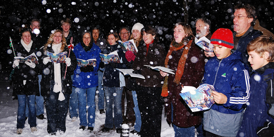 A semi-circle of carol singers standing in the snow holding their pages of carols. .