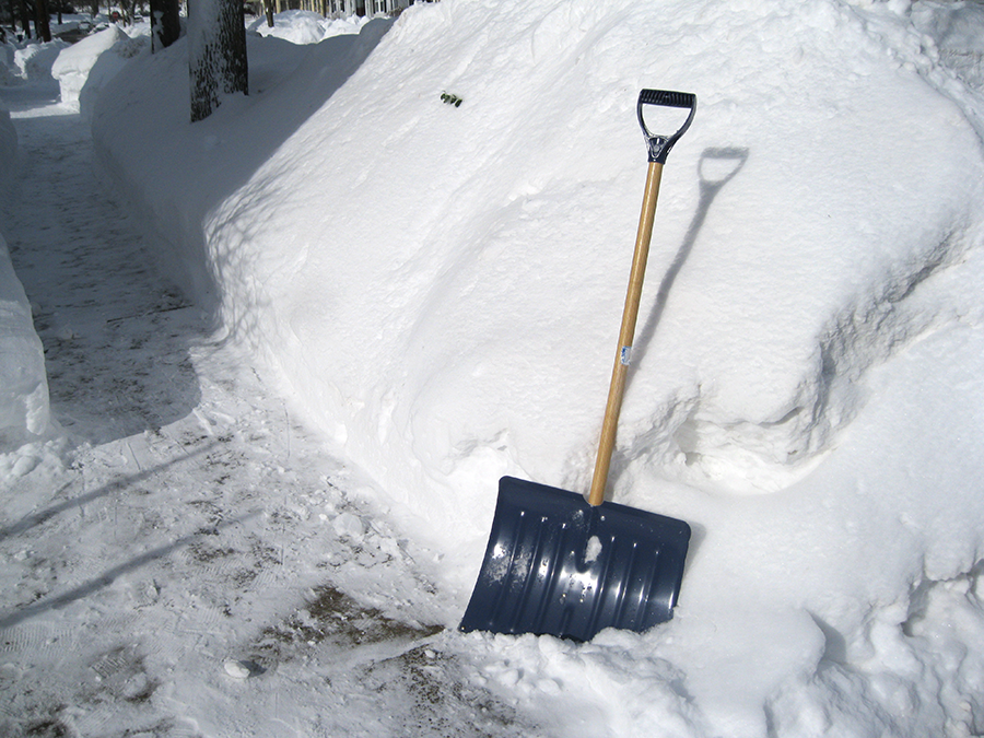 A snow shovel leans against a 6 foot wall of snow, with an icy sidewalk beside it.