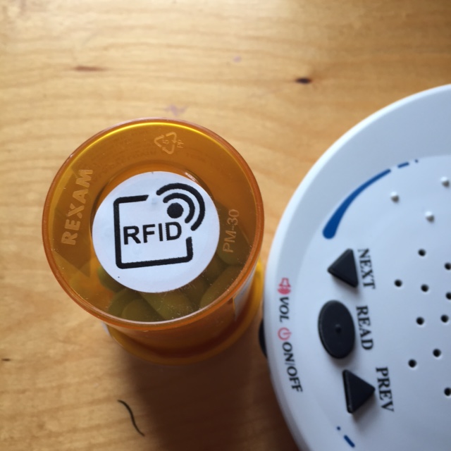 A ScripTalk label, called an RFID label, stuck to the bottom of a pill bottle. To the right there is the white curve and black control buttons on the ScripTalk Patient Station. 